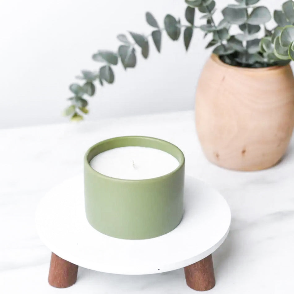 5 oz sage green ceramic candle. It is made from hemp + coconut wax and contains only clean and sustainable ingredients. the candle is resting on a white, wood footed pedestal with a vase with eucalyptus in the background.