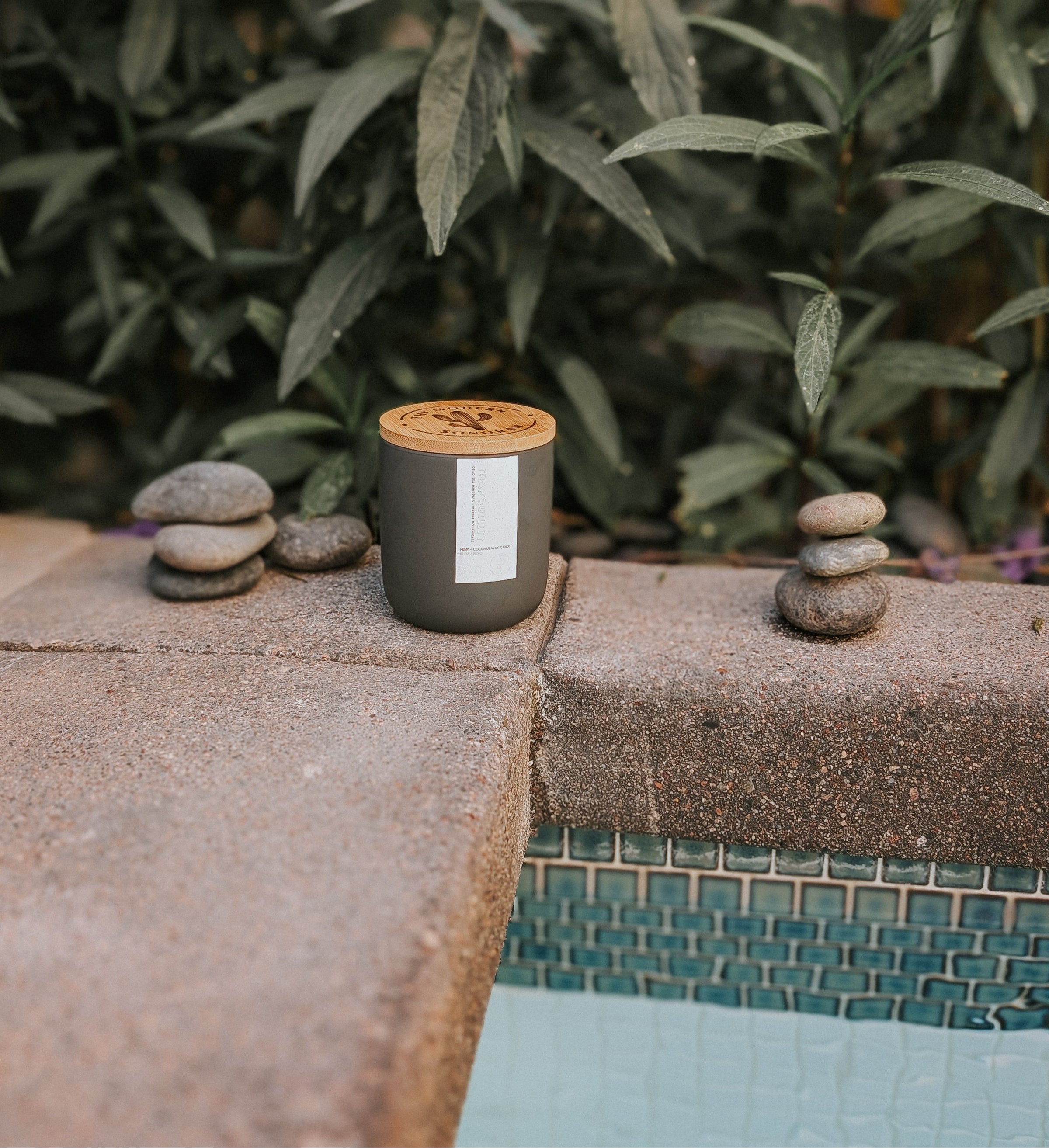 Black candle with wooden lid on a stone step surrounded by greenery.