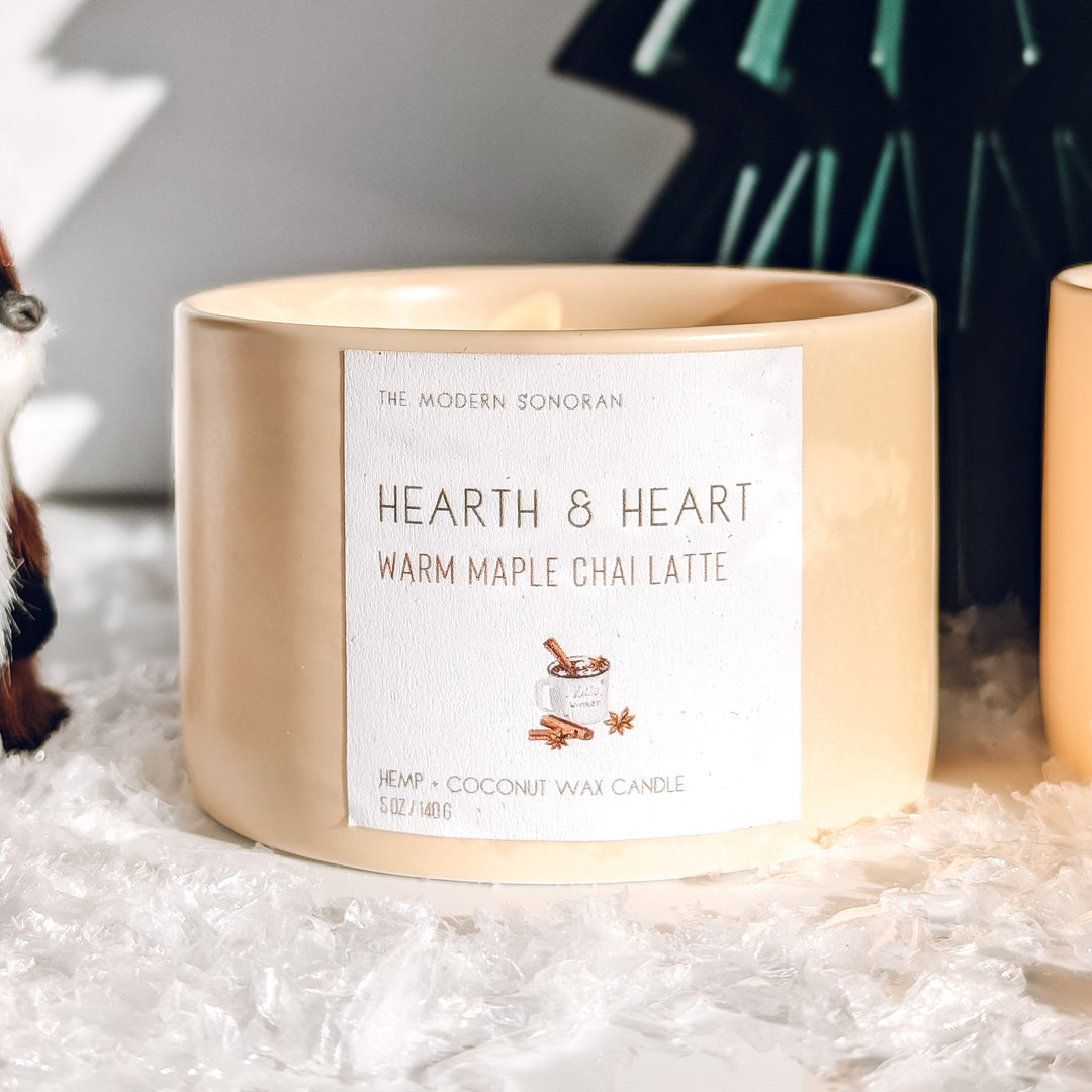 Candle labeled 'Hearth & Heart Warm Maple Chai Latte' with decorative trees and a bird figurine in the background.