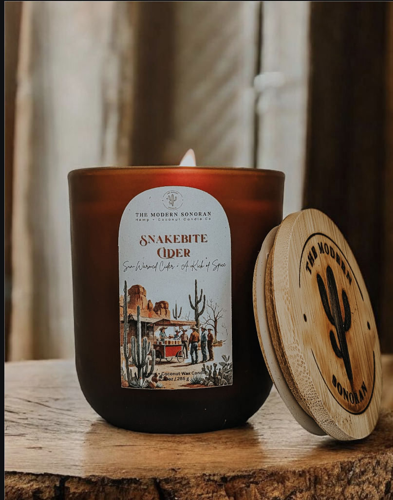 Fall candle with a label and wooden lid on a rustic surface. It's an apple cider candle called snakebite cider designed by the modern sonoran.