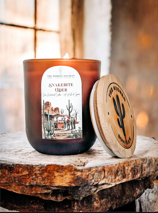 Southwestern candle called Snakebite Cider with a wooden lid on a rustic surface, featuring a label and wooden brand logo. 