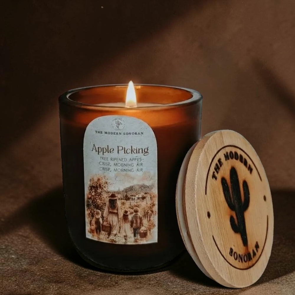 A lit amber glass candle jar with a white label reading 'Apple Picking' by The Modern Sonoran, shown beside a round natural wood lid engraved with a cactus and 'THE MODERN SONORAN'.