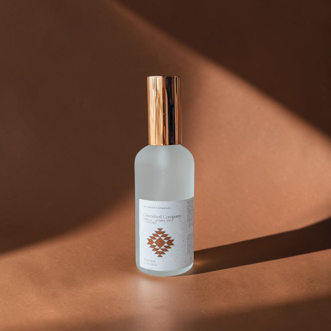 A frosted glass bottle of Cherished Company room spray with a metallic rose gold cap and a white label featuring a geometric pattern, set against a warm brown background with dramatic diagonal shadows.
