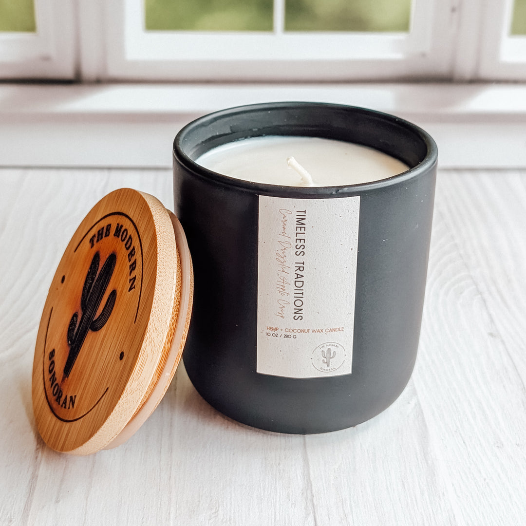 A close up shot of a Timeless Traditions candle in a black ceramic jar on a windowsill